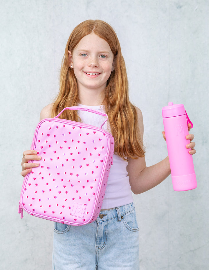 Girl holding a pink lunch bag with hearts on it and pink water bottle against a light gray background