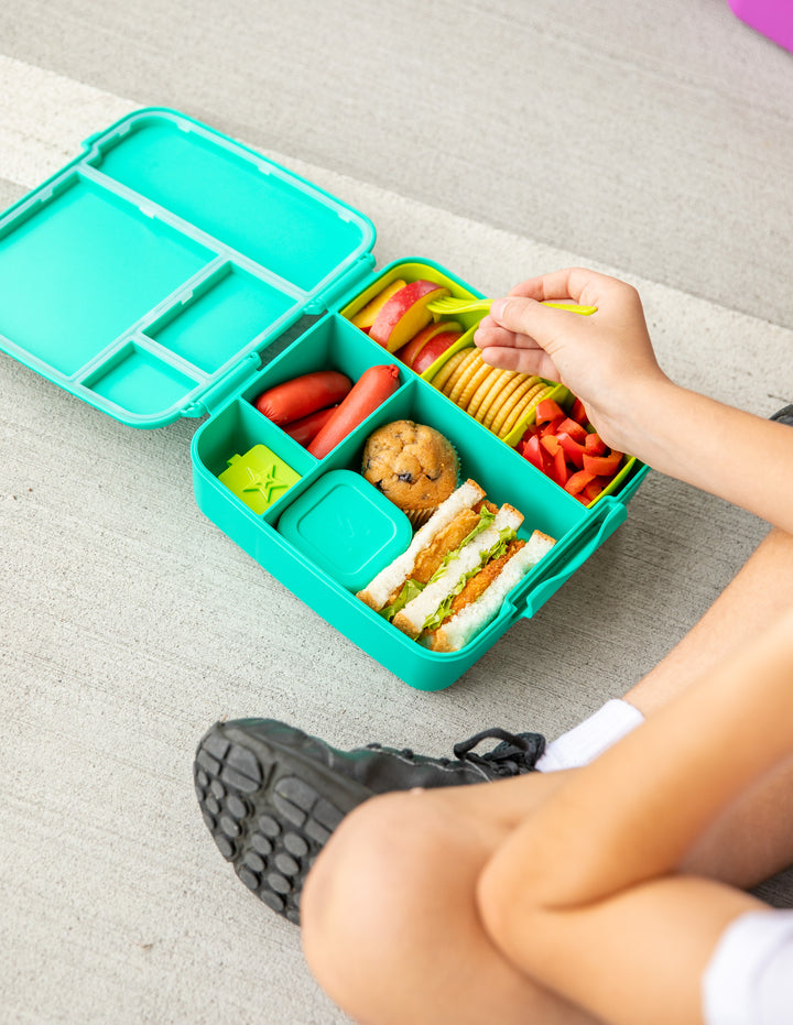 School child eating out of colourful bento lunch box with various food items 