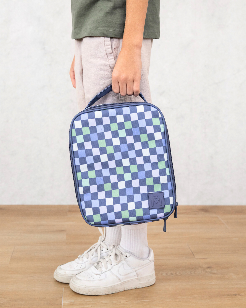 Person holding a blue and green checkered bag on a wooden floor with a white wall background