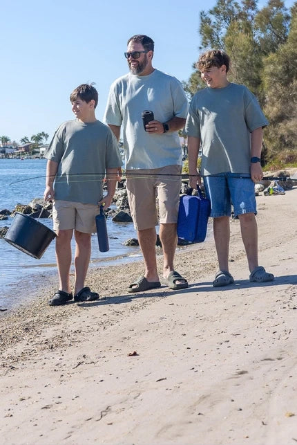 Three people walking on a sandy beach next to the ocean, wearing matching light gray t-shirts and casual shorts, with one person holding a black bucket and another holding a blue container.